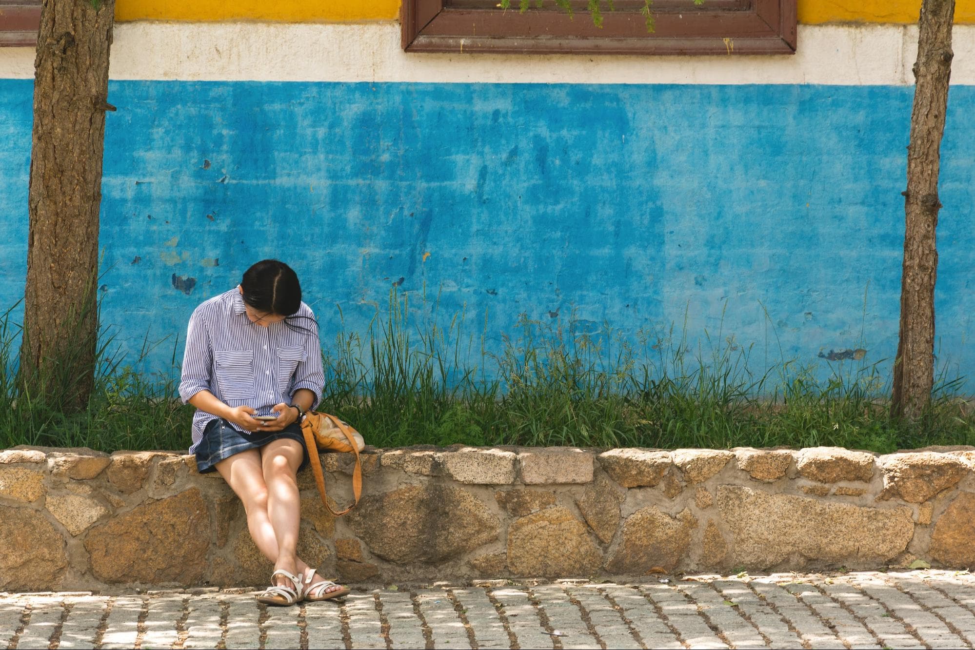 An image of a person perched on the left-hand side of a light brown stone wall in Argentina. Behind them is a wall that has been painted bright blue. On either side of the pictures are two painted tree trunks.