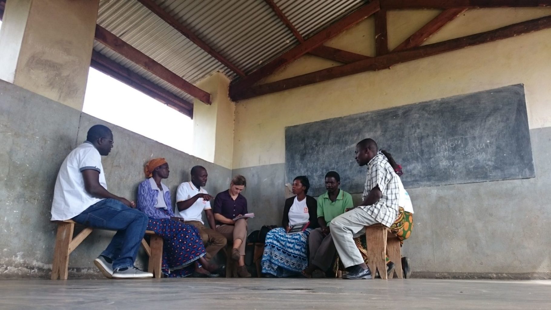 Welcoming three more partners to our Humanitarian Tech Support Programme an image of a group of people. They are sitting on chairs and are facing each other in a circle. They are having a discussion in a large room with a blackboard on the wall.