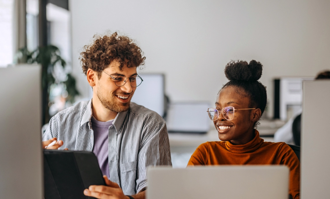 An image of two people sitting at a desk, looking at each other and smiling.