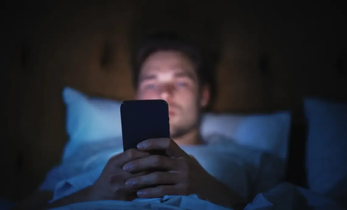 An image of a man sitting in a darkened room and using his mobile phone.