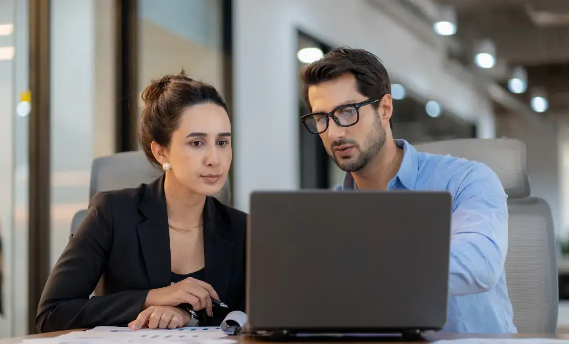 An image of a man and a women, sitting in an office setting and looking at a laptop.